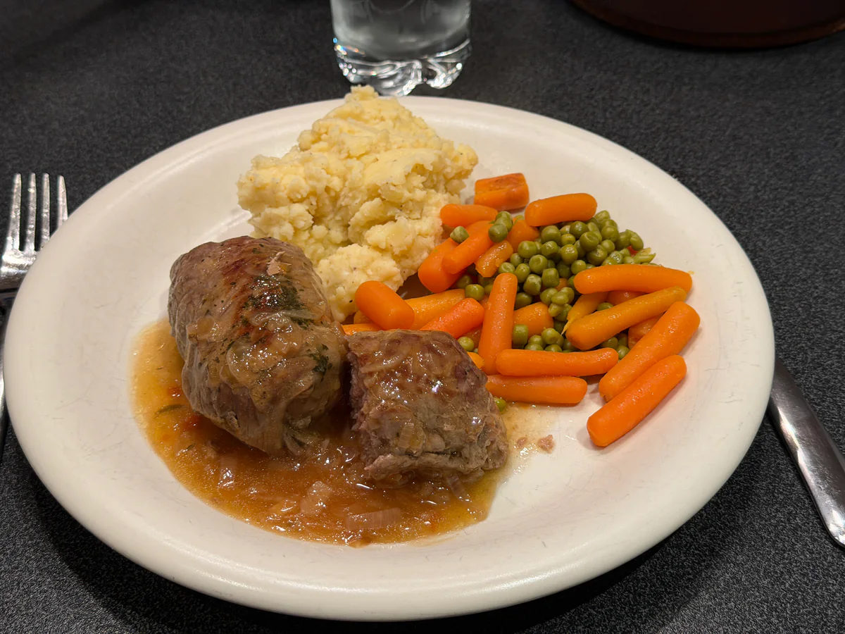 Vogels zonder kop met bruine uiensaus, puree, erwten en wortelen op een wit bord.