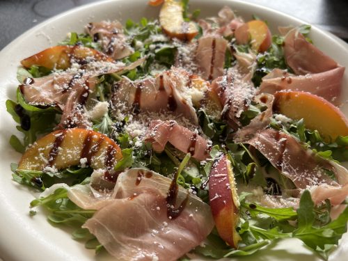en harmonie van prosciutto, verse perzik, en Parmezaan (of Feta) op een bedje van rucola, afgemaakt met een zoete balsamico crème.