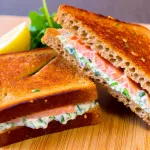 A Croque Norvégien sandwich, featuring two slices of toasted whole wheat bread. The sandwich is made with smoked salmon and herb cream cheese.