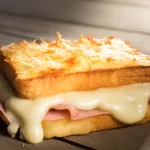 A close-up photo of a French-style Croque Monsieur sandwich with a layer of creamy béchamel sauce and melted grated cheese on top. The cheese layer is