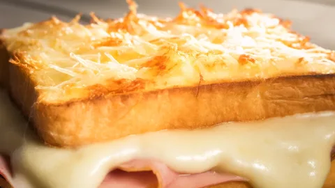 A close-up photo of a French-style Croque Monsieur sandwich with a layer of creamy béchamel sauce and melted grated cheese on top. The cheese layer is