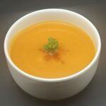 Kom warme wortelsoep met gember en kurkuma, afgewerkt met een takje peterselie en een vleugje cayennepoeder